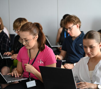 Studenci siedzą w sali i czytają informacje z laptopów.